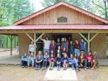 Students are gathered on the porch of the adobe building that has a red metal roof.